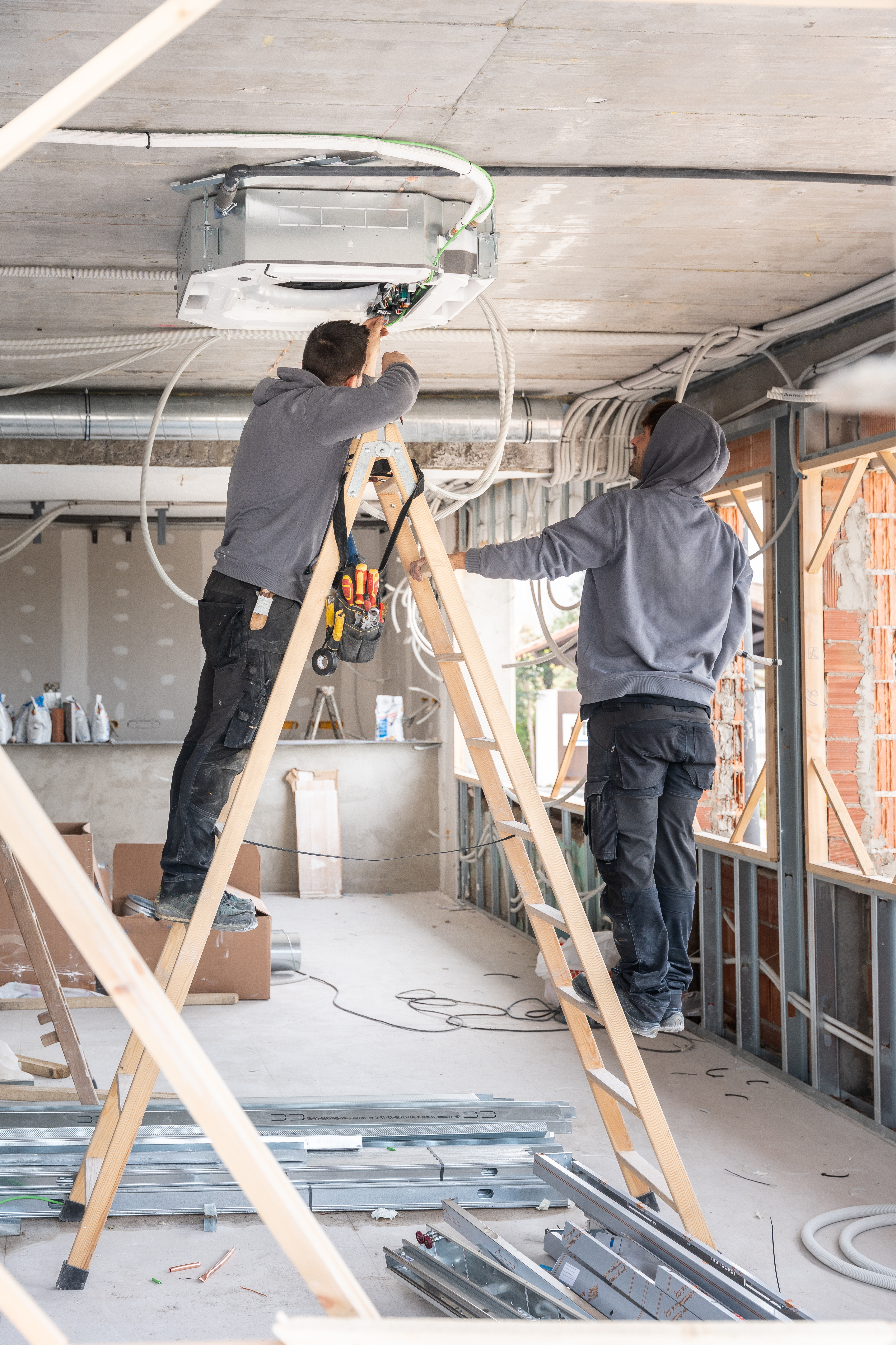 Two construction workers installing a new air conditioning unit in a building while working on ladders at a construction site, demonstrating teamwork and professional skill